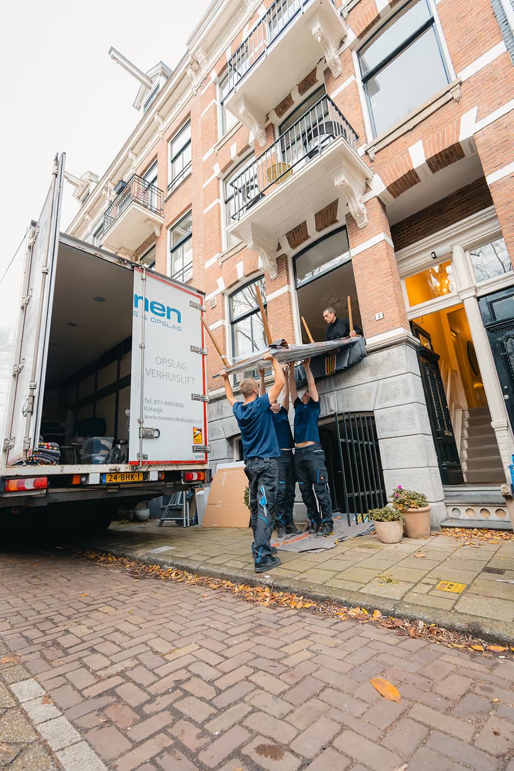 Verhuizers van Oomen tillen een ingepakt meubelstuk door het raam van een herenhuis naar buiten, naast de verhuiswagen